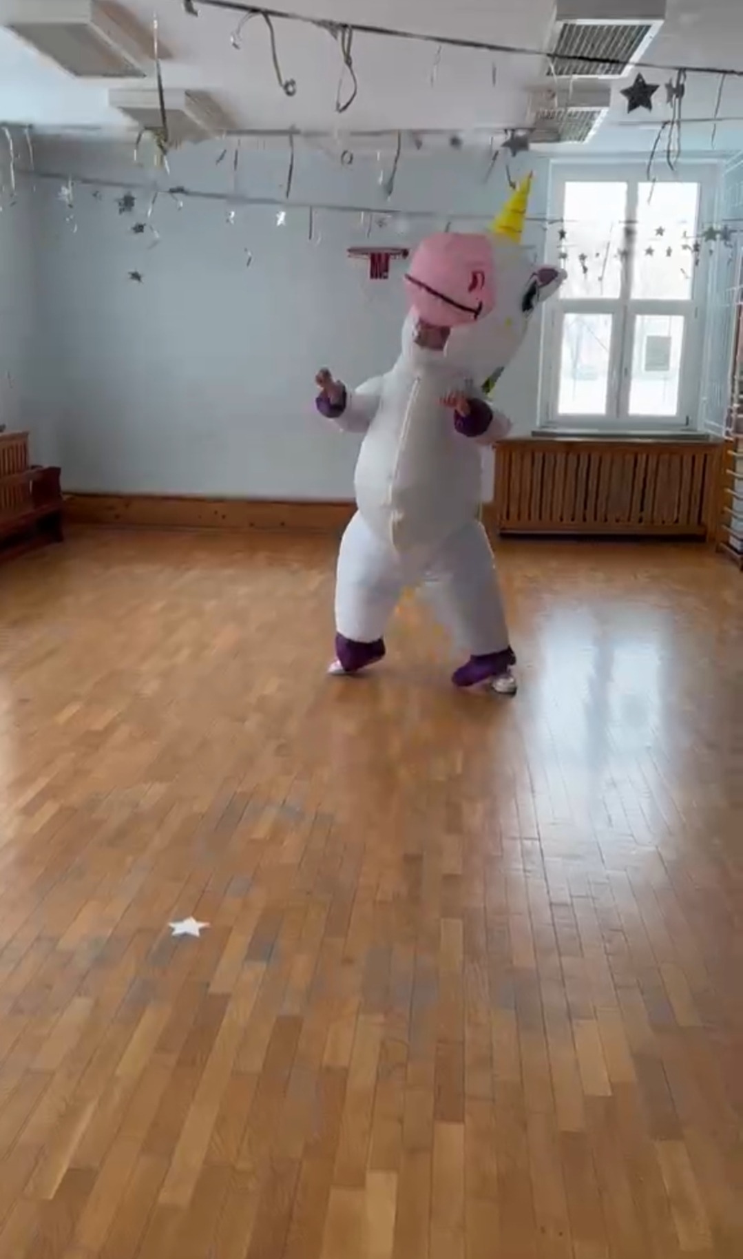 Animator w kostiumie jednorożca na parkiecie, dekoracje wiszą na suficie. Sala z oknami i ławkami. Kostium biały z różową głową i żółtym rogiem.