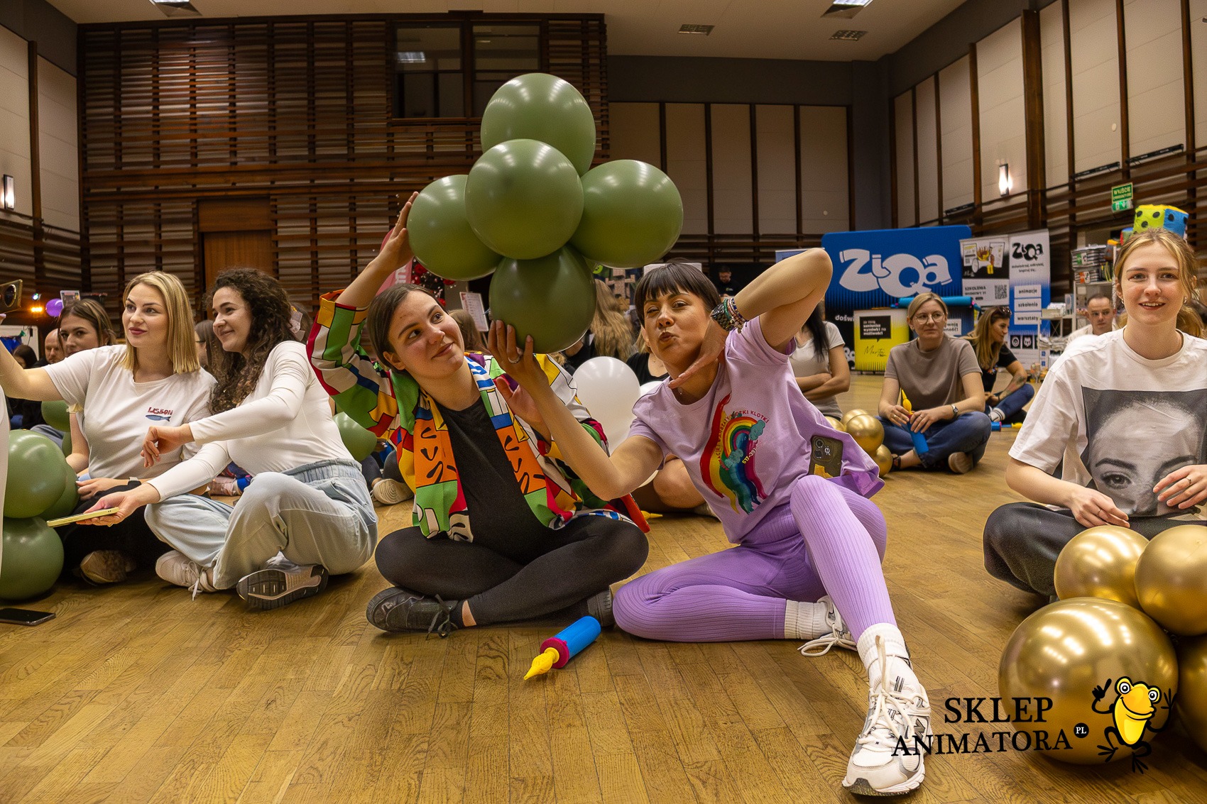 Grupa animatorów siedzi na podłodze z balonami, przygotowując się do imprezy. Widoczne kolorowe stroje i rekwizyty. W tle logo firmy eventowej.