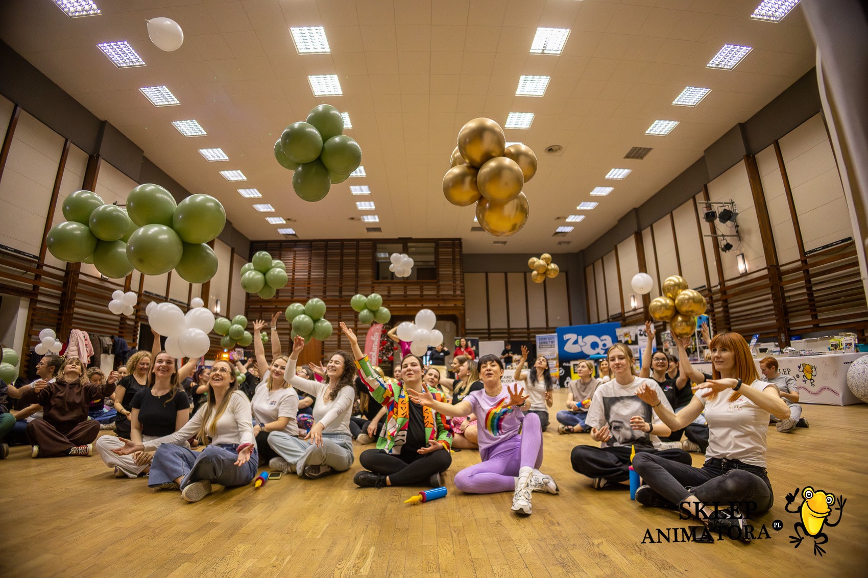 Grupa uśmiechniętych kobiet siedzi na podłodze sali gimnastycznej, unosząc ręce do góry w kierunku balonów w kolorach zieleni, złota i bieli. Impreza.