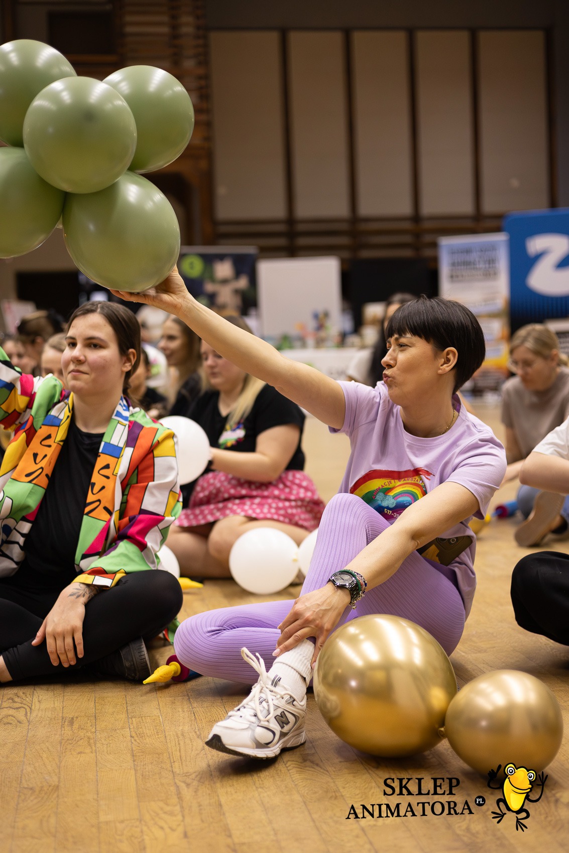 Animatorzy siedzą na podłodze z balonami: zielonymi, białymi i złotymi. Kobieta w fioletowym stroju trzyma zielone balony. Widoczny napis 'Sklep Animatora'.