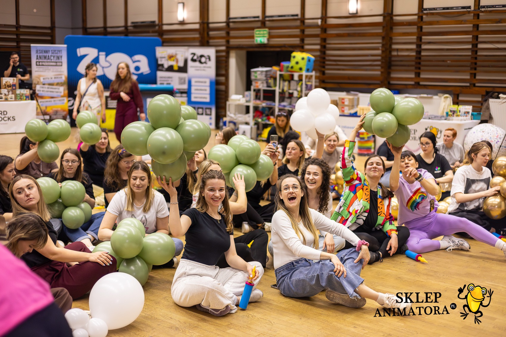 Grupa uśmiechniętych kobiet siedzi na podłodze z balonami w kolorze oliwkowym i białym, w tle banery reklamowe i regały z akcesoriami imprezowymi.