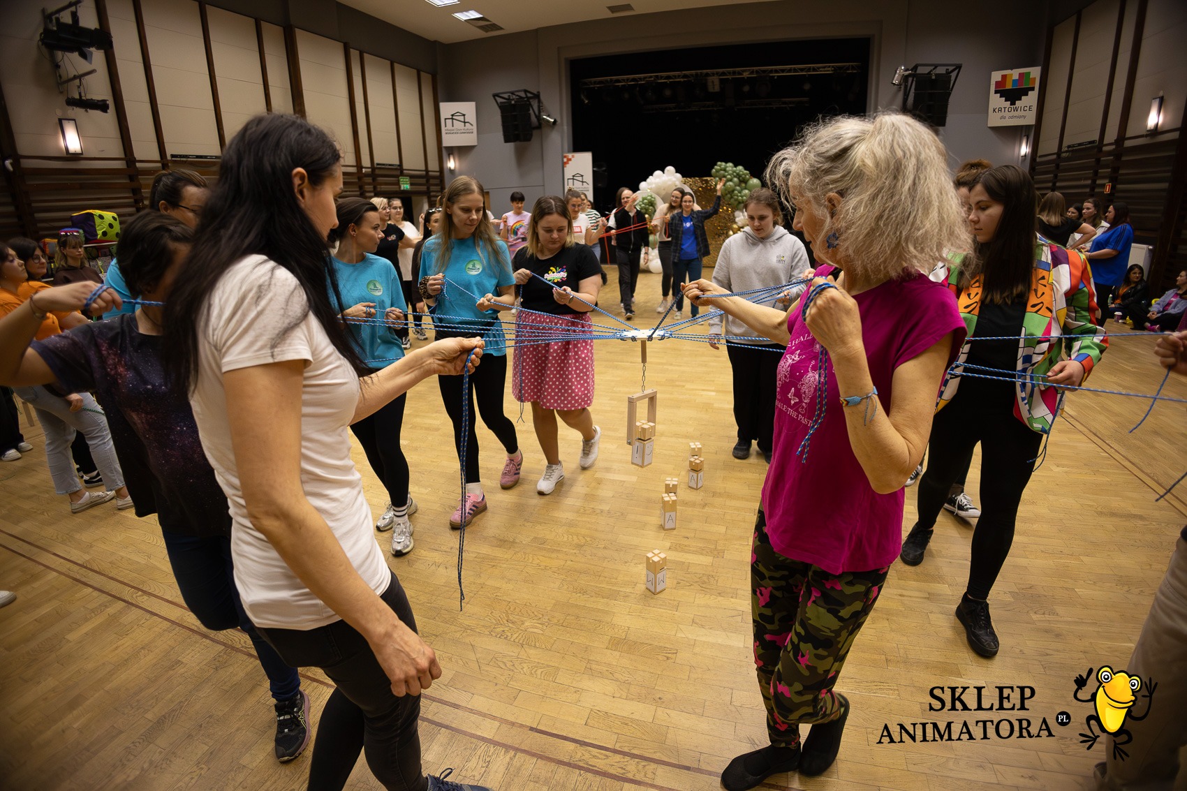 Grupowa zabawa z linami i drewnianymi klockami w sali gimnastycznej. Uczestnicy trzymają liny, tworząc skomplikowaną sieć. W tle widoczna scena i dekoracje.