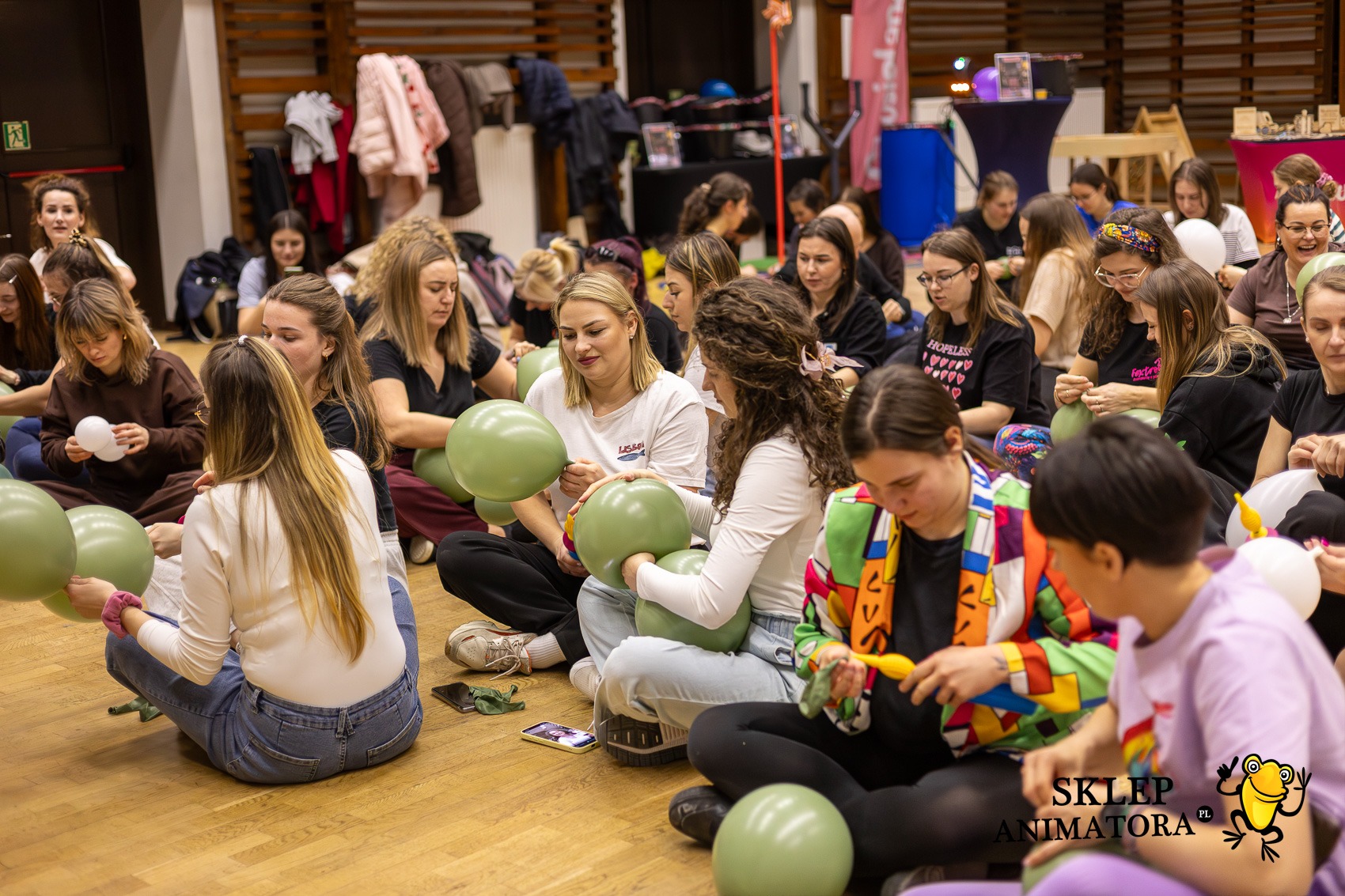 Warsztaty z animacji: grupa kobiet siedzi na podłodze, dmuchając zielone balony. W tle widoczne dekoracje i sprzęt eventowy. Jasne oświetlenie, parkiet.