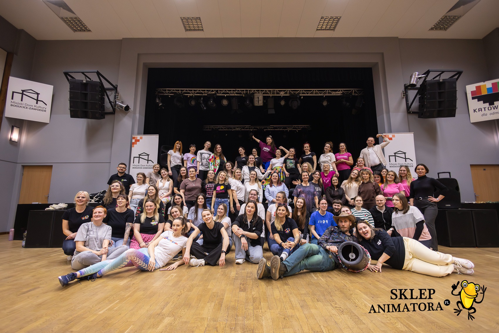 Grupowe zdjęcie zespołu animatorów na scenie z logo 'Sklep Animatora' w prawym dolnym rogu. W tle scena i banery Miejskiego Domu Kultury.