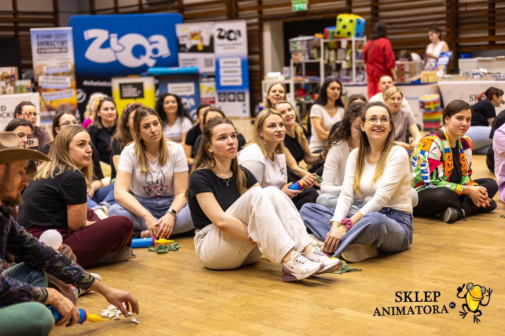 Grupa osób siedzi na podłodze podczas szkolenia animatorów, w tle materiały reklamowe i rekwizyty. Widoczny fragment logo 'Sklep Animatora'.