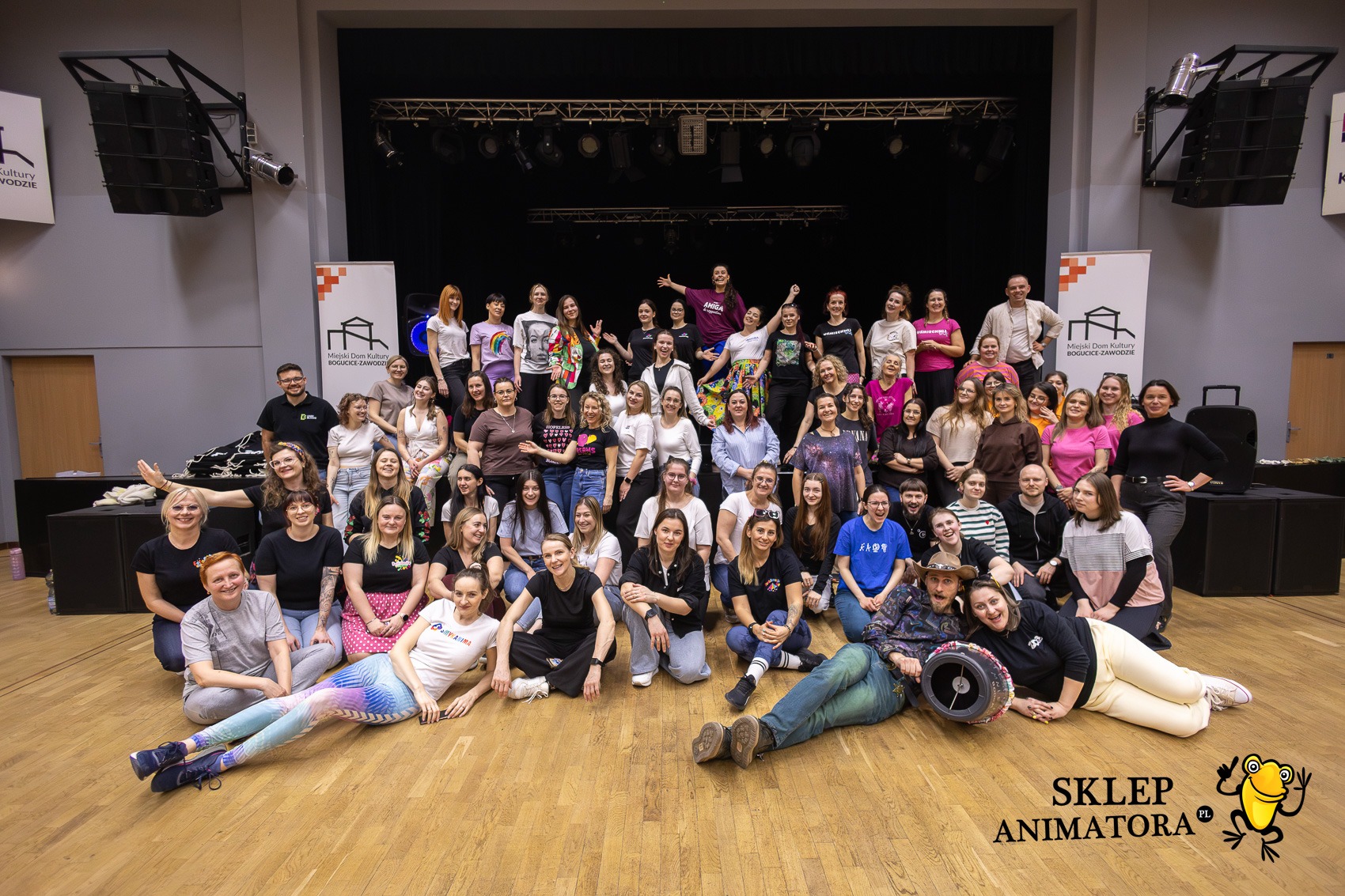 Grupowe zdjęcie zespołu animatorów na scenie z nagłośnieniem i oświetleniem, z logo 'Sklep Animatora' w prawym dolnym rogu. Uśmiechnięte twarze, różnorodne stroje.