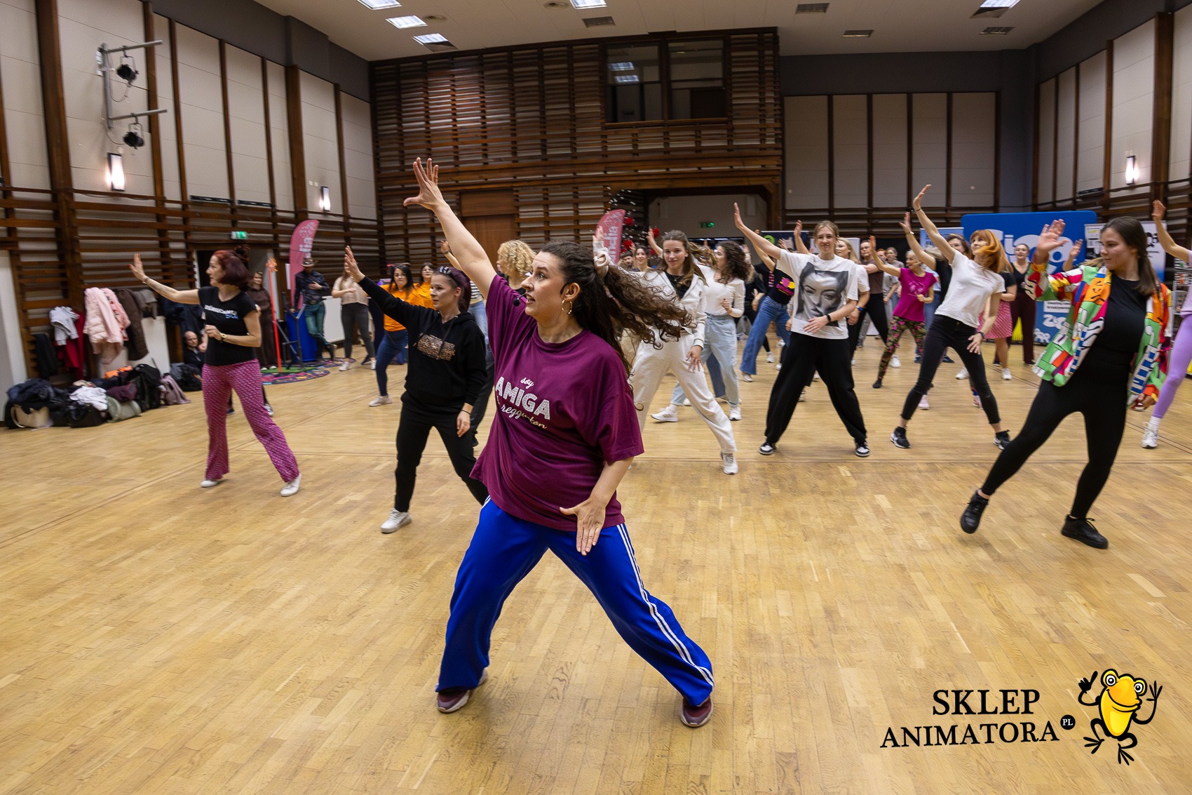 Grupa kobiet w różnym wieku tańczy w sali gimnastycznej, prowadzona przez instruktorkę w fioletowej koszulce i niebieskich spodniach. Parkiet, wysokie okna, logo animatora.