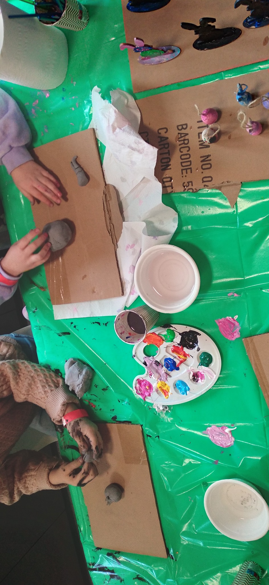 Warsztaty plastyczne dla dzieci: umazane farbą ręce formują glinę na kartonie, obok paleta z farbami i inne materiały plastyczne na zielonym stole.
