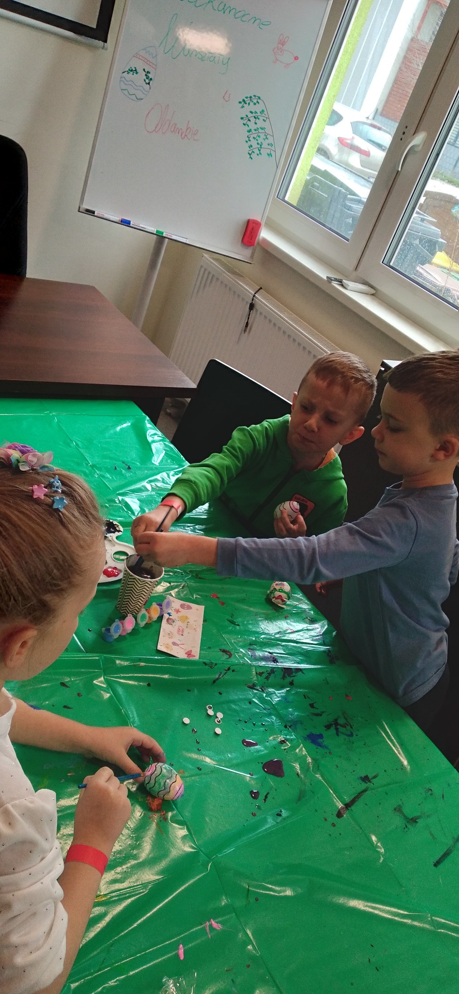 Dzieci malują pisanki na zielonym stole, widoczne farby, pędzle i dekoracje. Na tablicy rysunki wielkanocne. Kreatywne warsztaty dla dzieci.