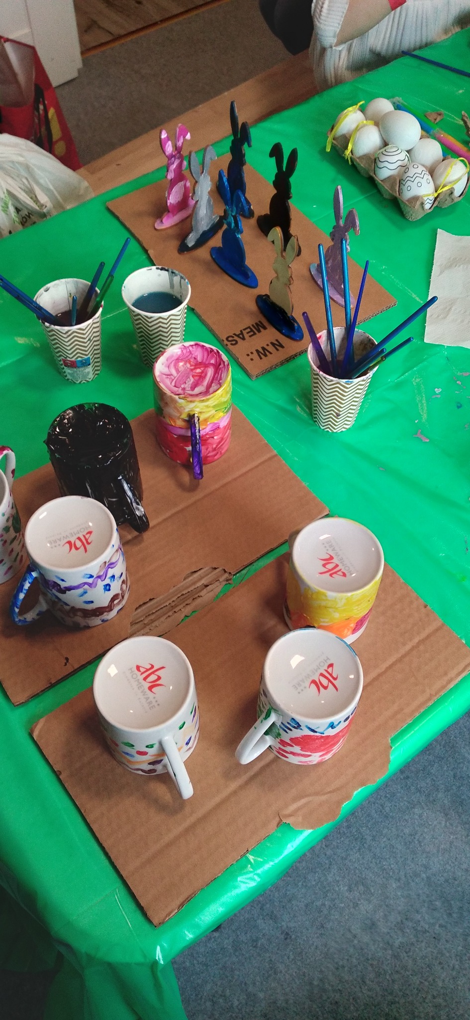 Warsztat malowania: kubki, jajka i drewniane zajączki na zielonym stole, pędzle w kubkach. Kreatywne zajęcie dla dzieci, rękodzieło.