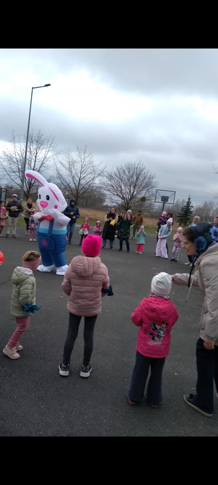 Animator w stroju dmuchanego królika bawi dzieci na zewnątrz. Uczestnicy w różowych kurtkach, pochmurne niebo w tle. Zabawa na świeżym powietrzu.