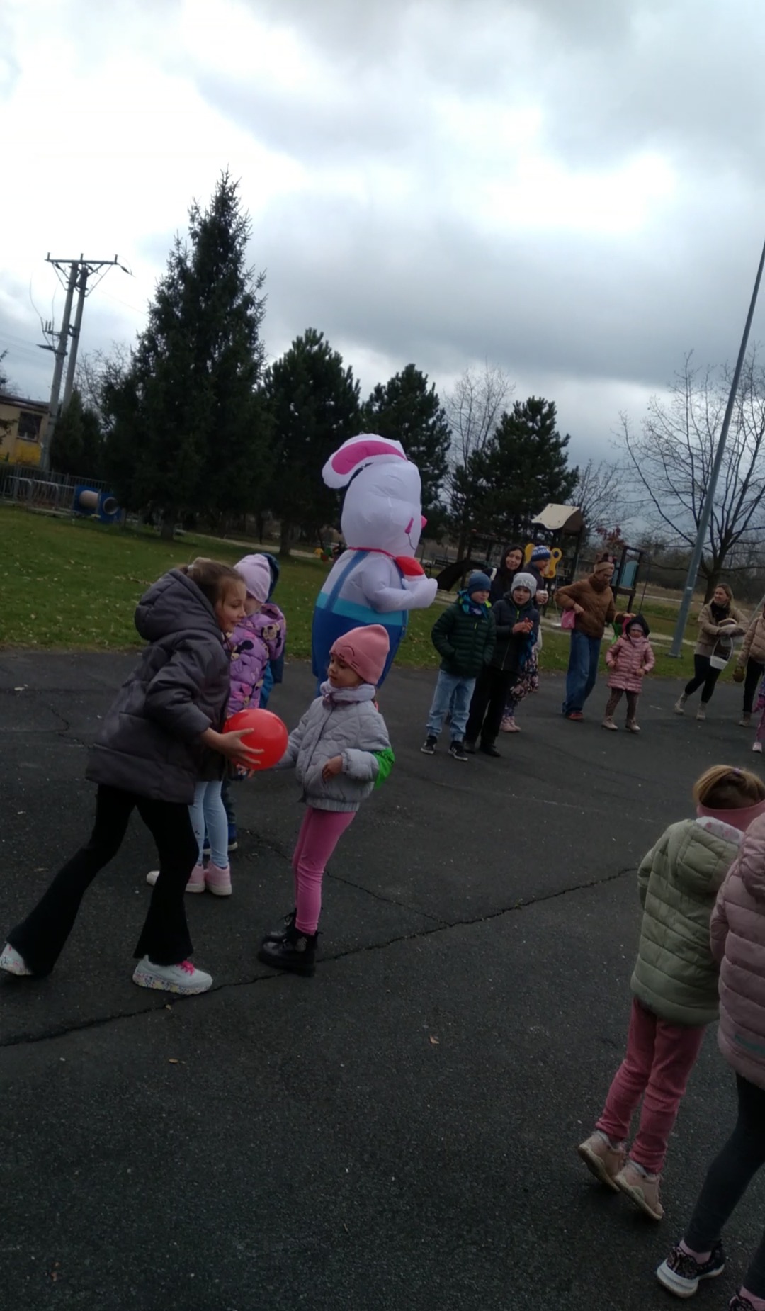Animator w kostiumie królika bawi dzieci na zewnątrz, w tle grupa rodziców i pochmurne niebo. Zabawa z czerwonym balonem.