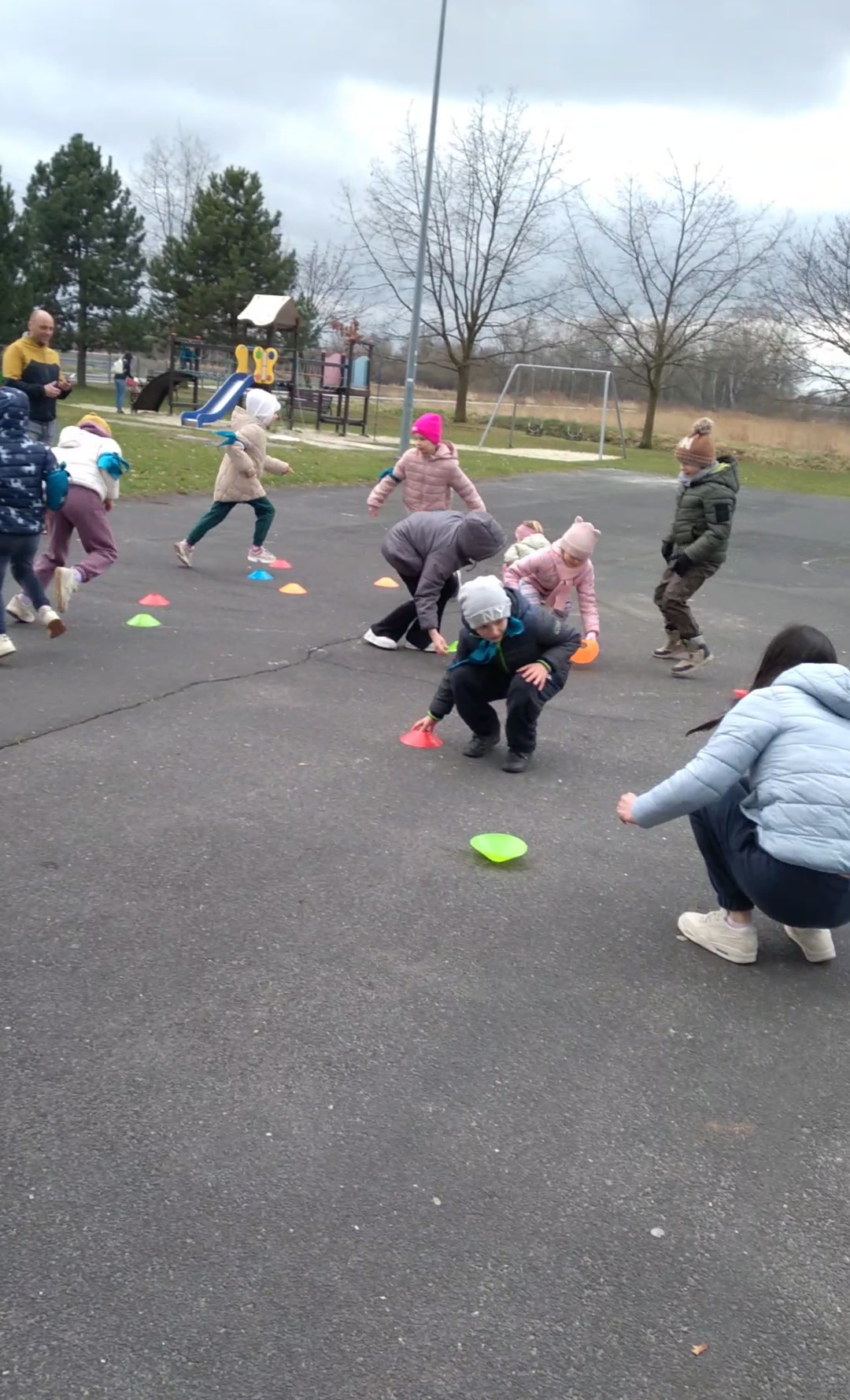 Grupka dzieci w różnokolorowych kurtkach i czapkach bawi się na zewnątrz, ustawiając kolorowe pachołki na szarym asfalcie. W tle plac zabaw i drzewa.