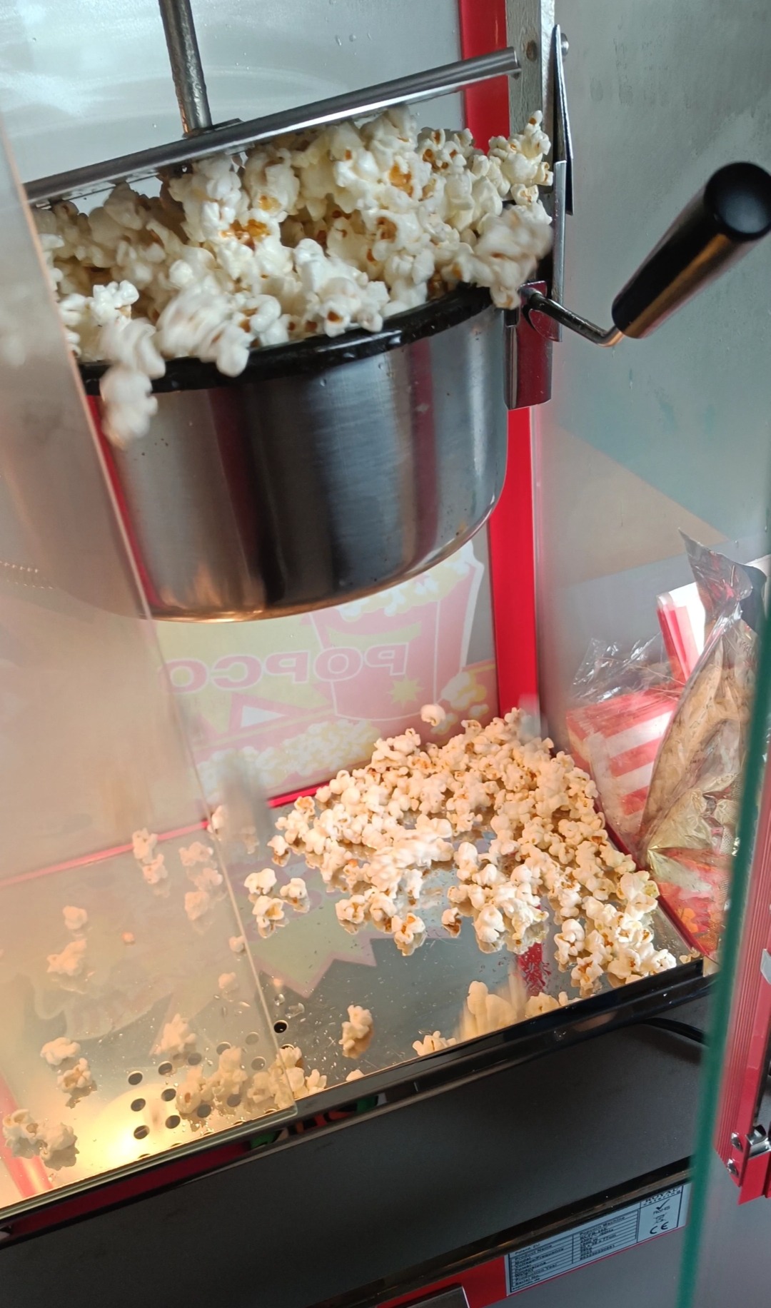 Automat do popcornu w trakcie pracy, świeżo uprażony popcorn wysypuje się na tacę. Widoczne opakowania na popcorn w tle.