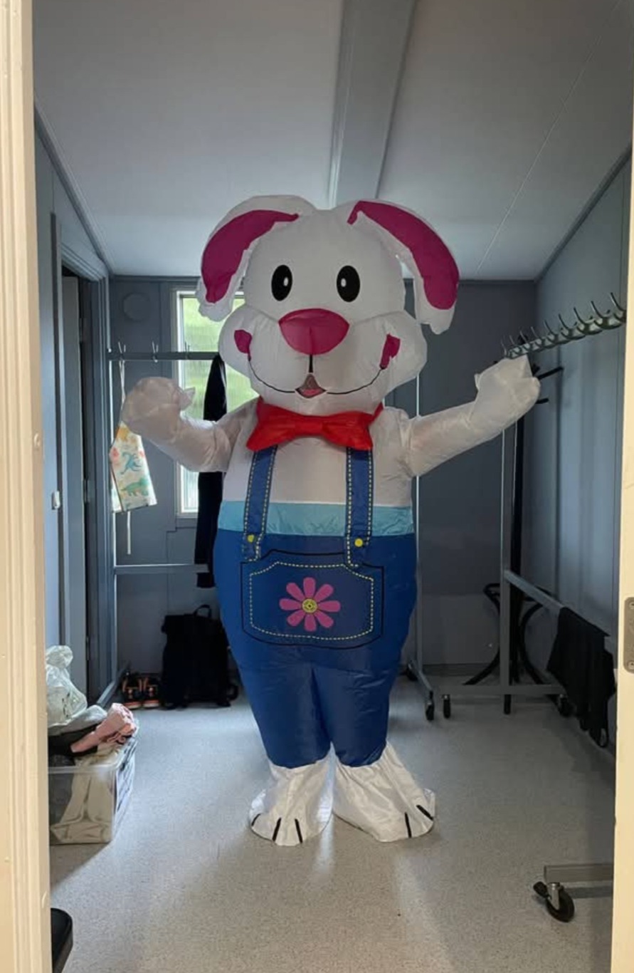 Nadmuchiwany królik w niebieskich ogrodniczkach z różowym kwiatkiem, czerwona mucha, białe futro i różowe uszy stoi w pomieszczeniu. W tle wieszaki i okno.