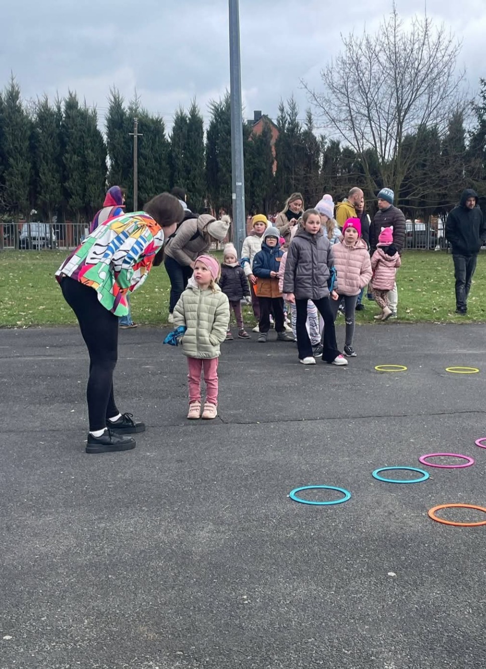 Animator w kolorowej kurtce prowadzi zajęcia z dziećmi na zewnątrz, wykorzystując kolorowe obręcze. Dzieci ubrane w zimowe kurtki obserwują uważnie.