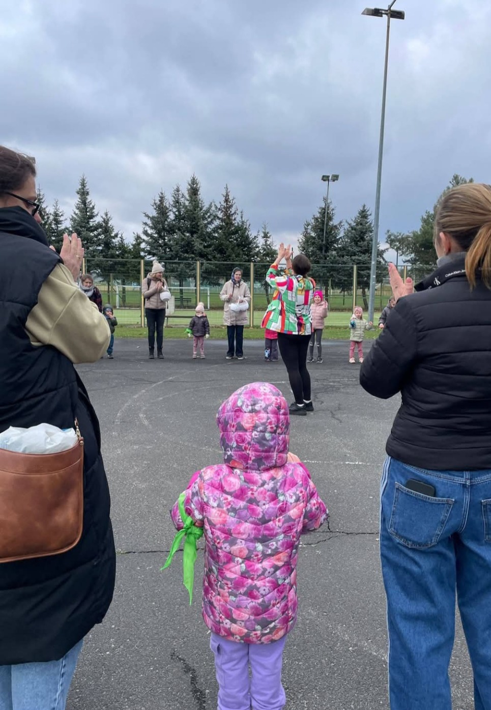 Animator w kolorowej kurtce prowadzi grupę dzieci i dorosłych na zewnątrz, wszyscy klaszczą. Widok zza pleców dziecka w różowej kurtce.