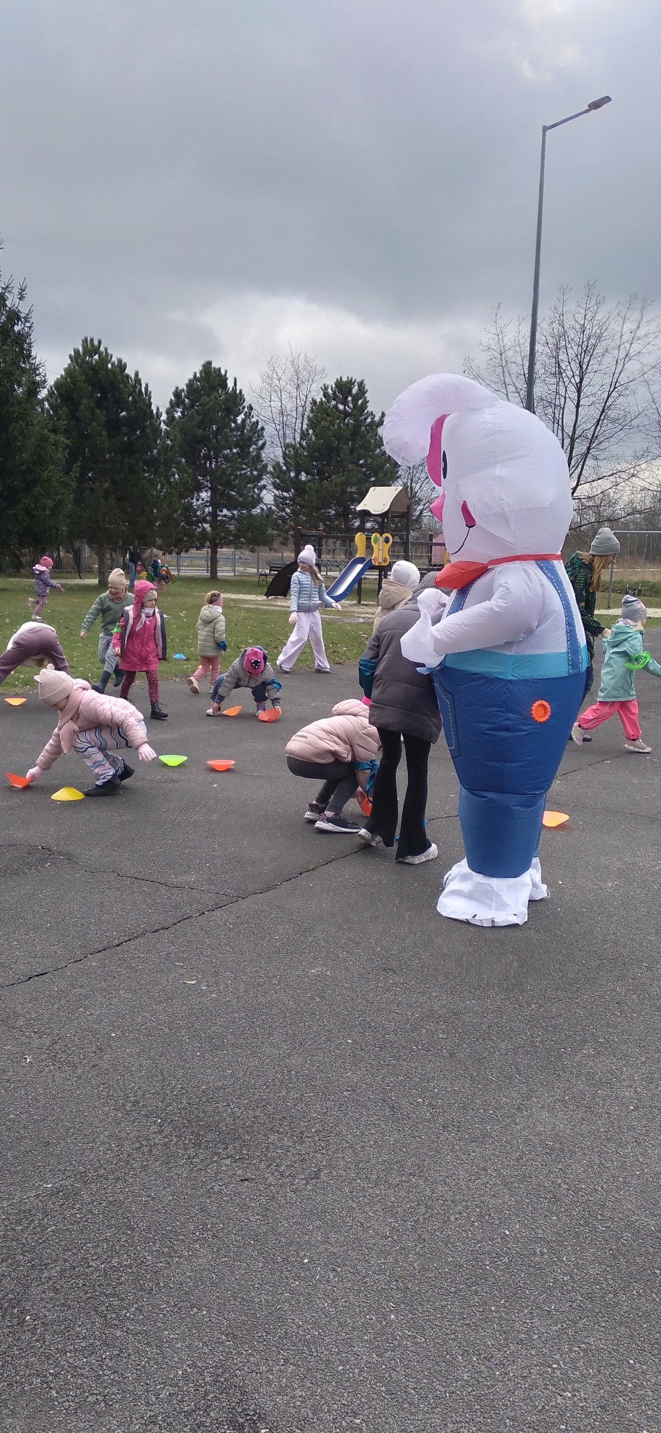 Animator w stroju dmuchanego królika prowadzi zabawy na zewnątrz dla grupy dzieci, z kolorowymi pachołkami na asfalcie i placem zabaw w tle.
