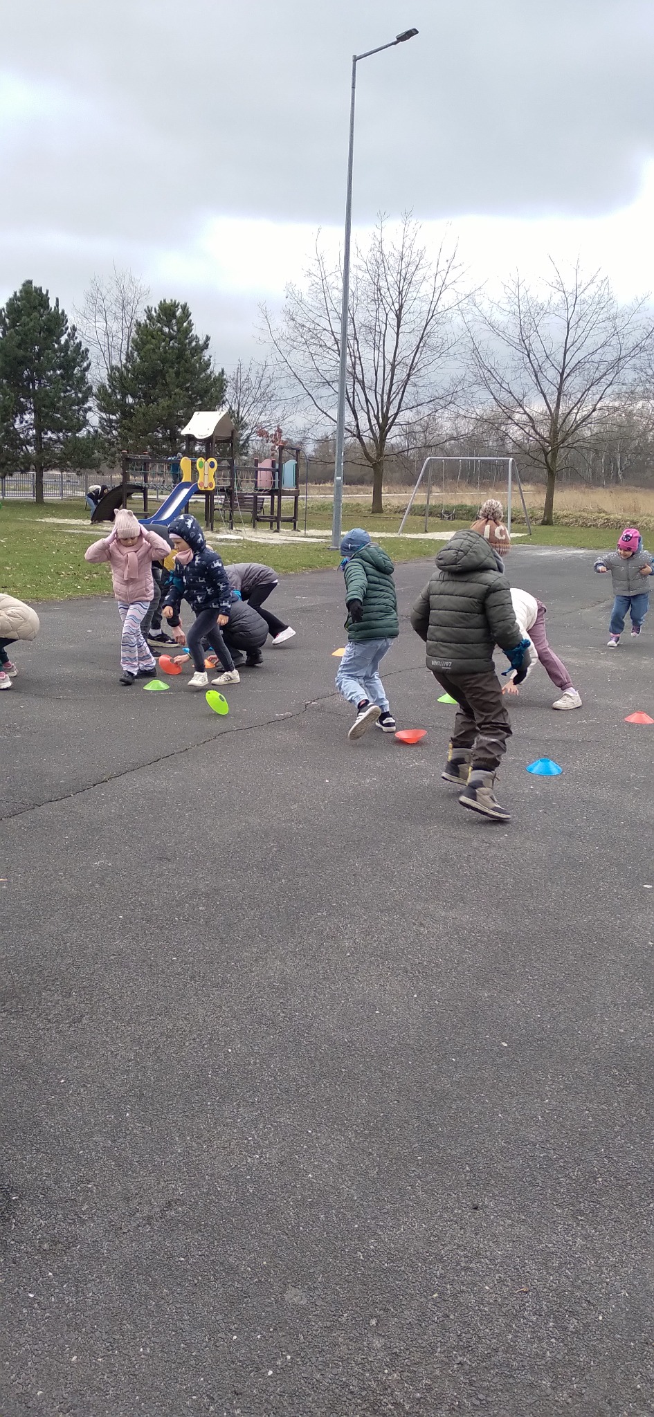 Grupa dzieci w kurtkach bawi się na asfalcie, omijając kolorowe pachołki. W tle plac zabaw i drzewa bez liści, pochmurne niebo. Aktywność na świeżym powietrzu.