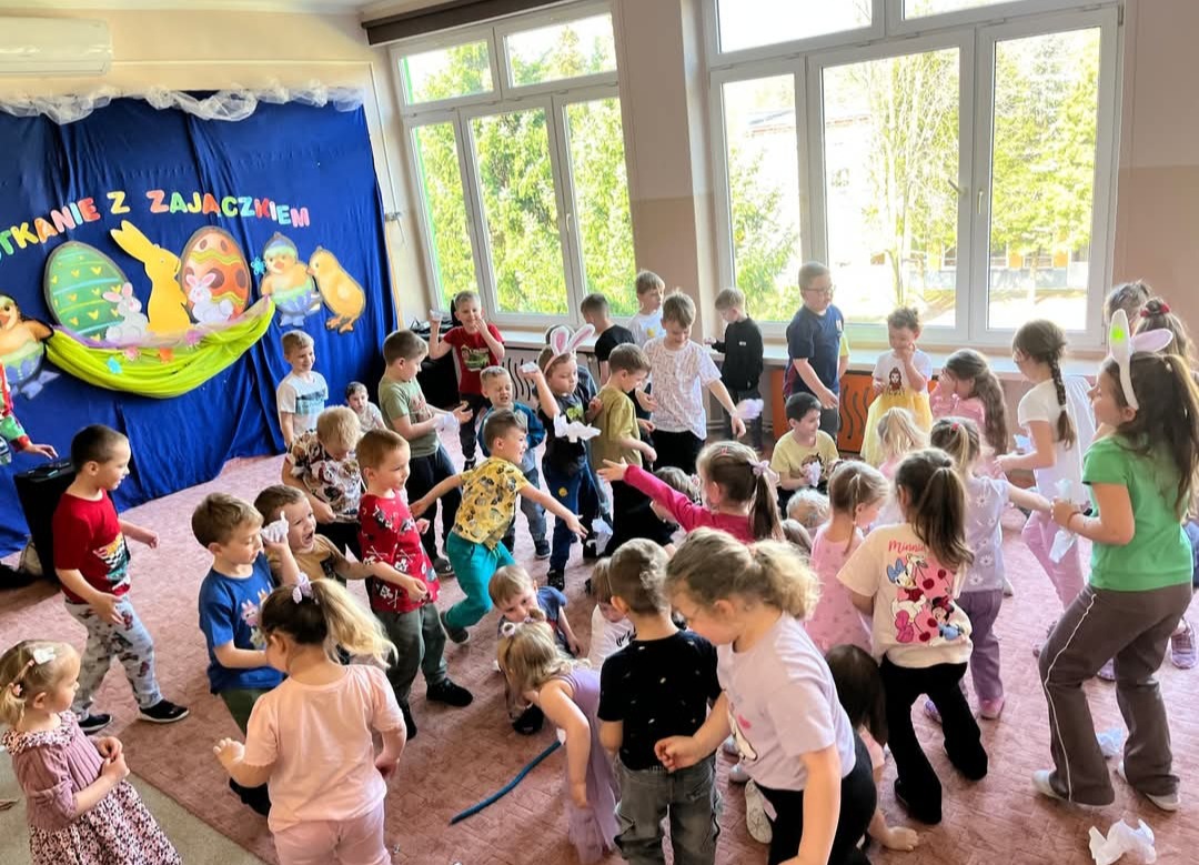 Radosne dzieci w sali podczas zabawy z okazji Wielkanocy, dekoracje z zajączkami i pisankami w tle, jasne wnętrze, dynamiczne ujęcie.