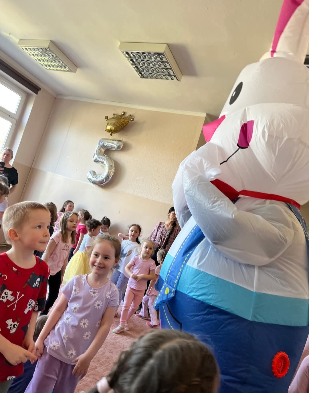 Dzieci obserwują dmuchanego królika podczas urodzin. W tle balon w kształcie cyfry 5 i korony. Sala oświetlona lampami. Uśmiechnięte dzieci w różnym wieku.