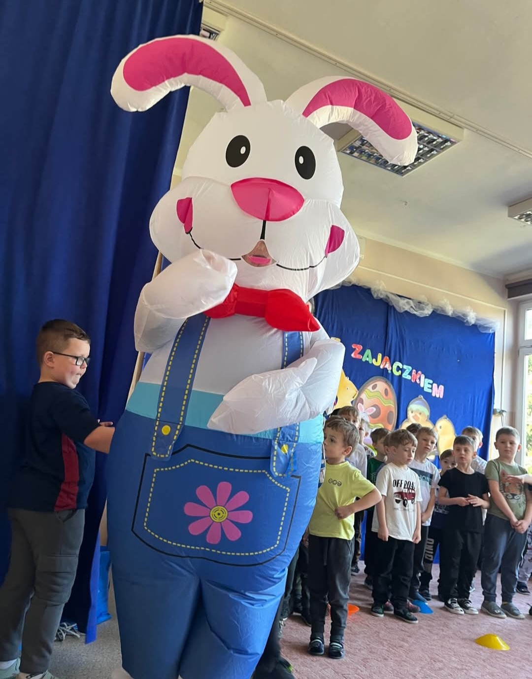 Animator w kostiumie dmuchanego zająca wielkanocnego w otoczeniu dzieci podczas wydarzenia. Dekoracja z napisem 'Zajączkiem'.
