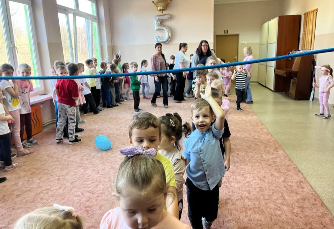 Dzieci ustawione w rzędzie, bawiące się pod liną podczas imprezy urodzinowej z balonem w tle, prowadzone przez animatorów. W tle balon w kształcie cyfry 5.