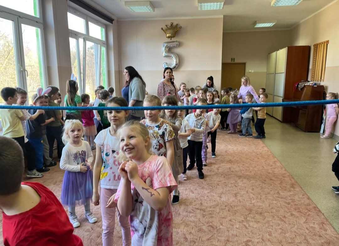 Uśmiechnięte dzieci ustawione w rzędzie, prowadzone przez animatora z liną, w tle balon w kształcie cyfry 5, sala udekorowana na przyjęcie urodzinowe.