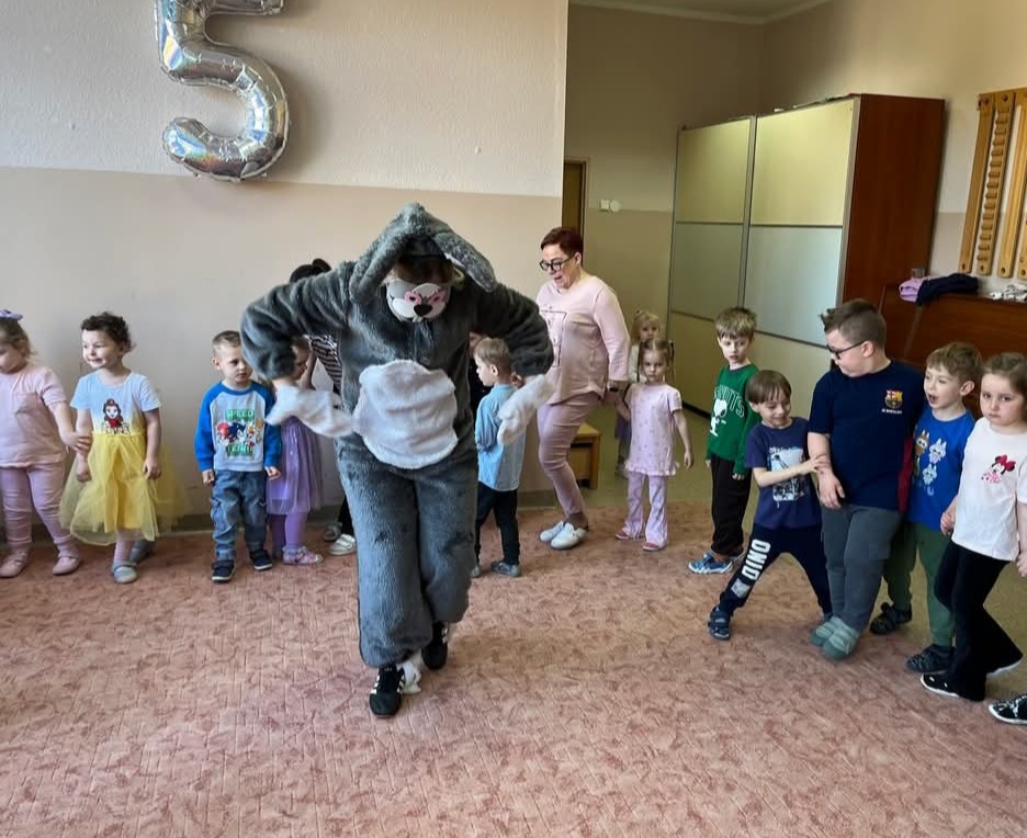 Animator w kostiumie królika bawi grupę dzieci w sali, balon w kształcie cyfry 5 w tle. Dzieci ubrane w kolorowe stroje, animator w szarym kostiumie.