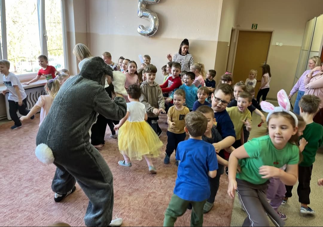 Animator w kostiumie królika bawi grupę roześmianych dzieci w sali z balonem w kształcie cyfry 5. Dzieci ubrane w kolorowe stroje, niektóre z uszami królika.