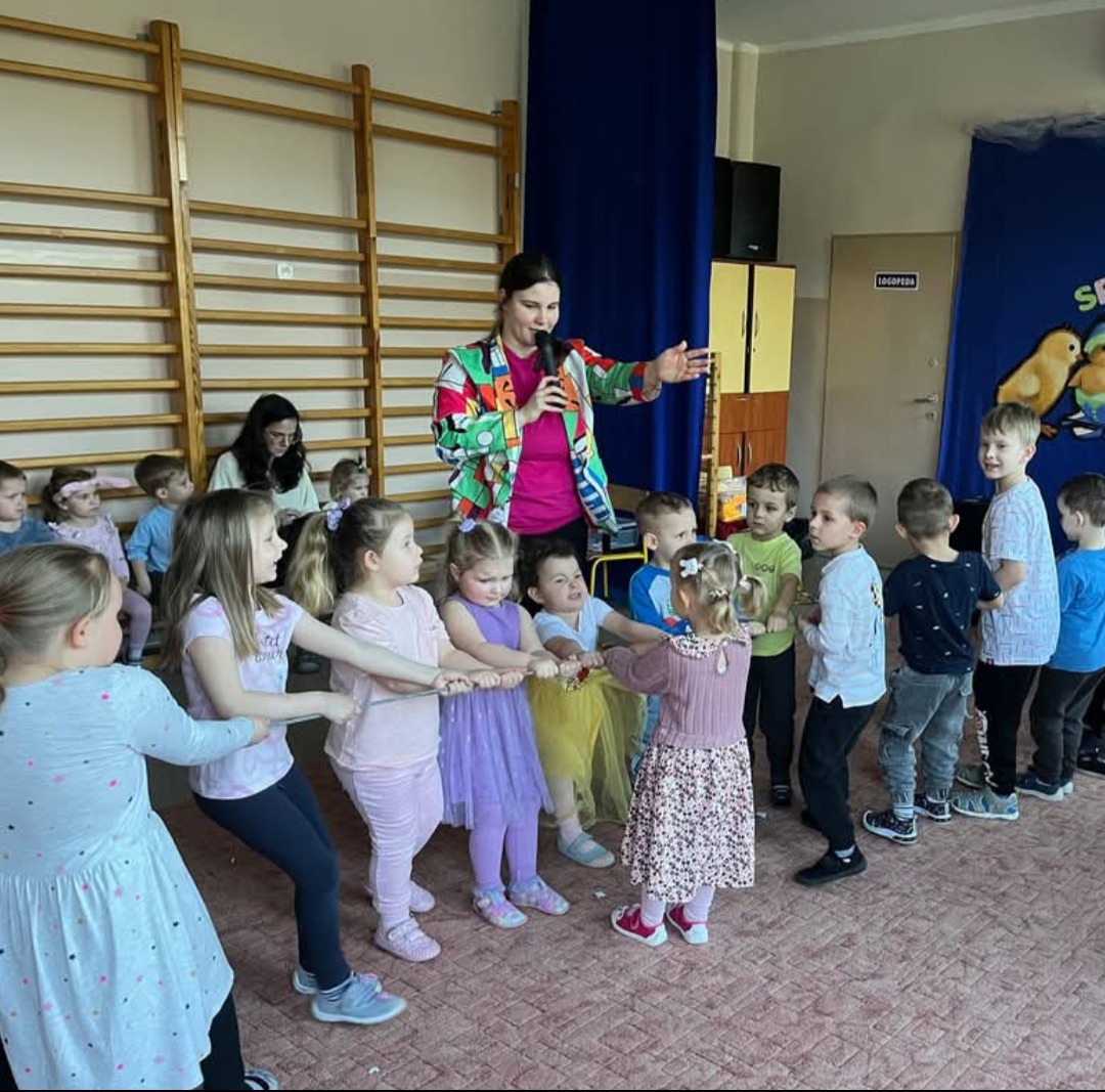 Animator w kolorowej marynarce prowadzi zabawę w przeciąganie liny dla grupy dzieci w sali. W tle widoczna ściana z dekoracjami.