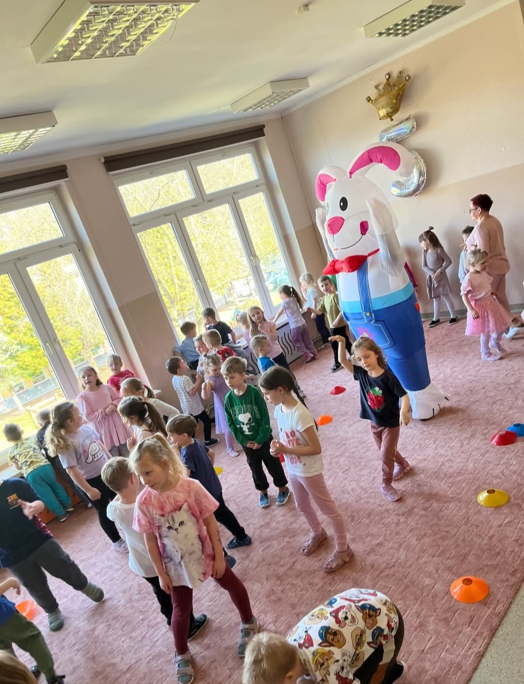 Sala zabaw wypełniona dziećmi i animatorem w stroju królika. Balon w kształcie korony i cyfry 5 w tle. Dzieci w różnym wieku, wesoła atmosfera.
