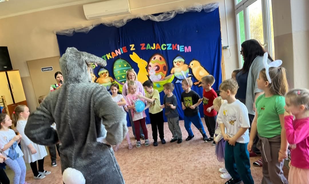 Spotkanie z Zajączkiem: animator w kostiumie zająca prowadzi zabawy dla dzieci w przedszkolu, na tle dekoracji wielkanocnych. Dzieci ustawione w kręgu.