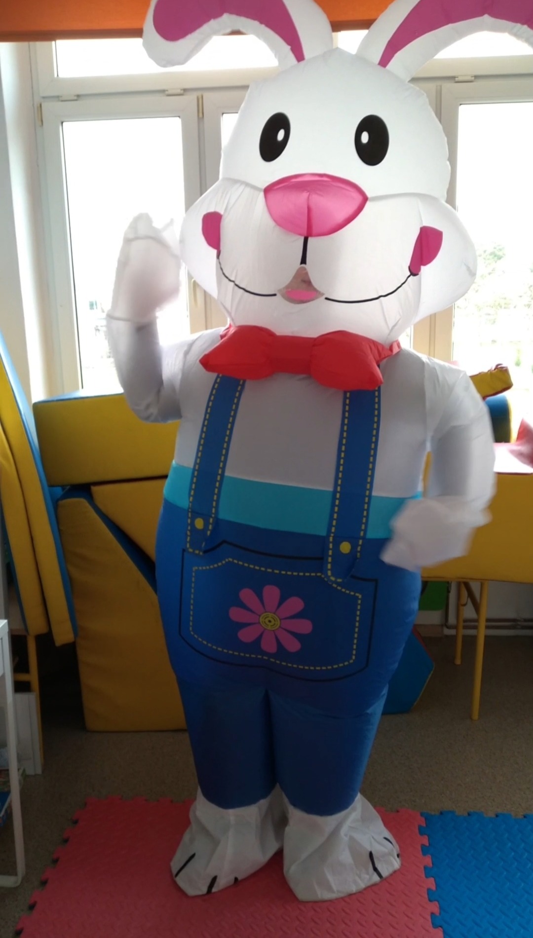 Nadmuchiwany kostium królika w niebieskim kombinezonie z różowym kwiatem, czerwona muszka, białe rękawice, na tle okna i kolorowych mebli.