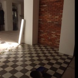 Byd-Rem - Ułożona podłoga w szachownicę, częściowo oświetlona lampą budowlaną, z widocznymi elementami poziomowania i wiadrem farby, w tle ściana z cegły w łuku.