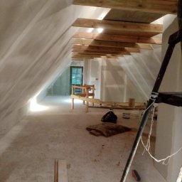 Byd-Rem - Poddasze w trakcie prac wykończeniowych: szpachlowane ściany, drewniana konstrukcja sufitu, drabina malarska w prawym dolnym rogu, okno w głębi pomieszczenia.