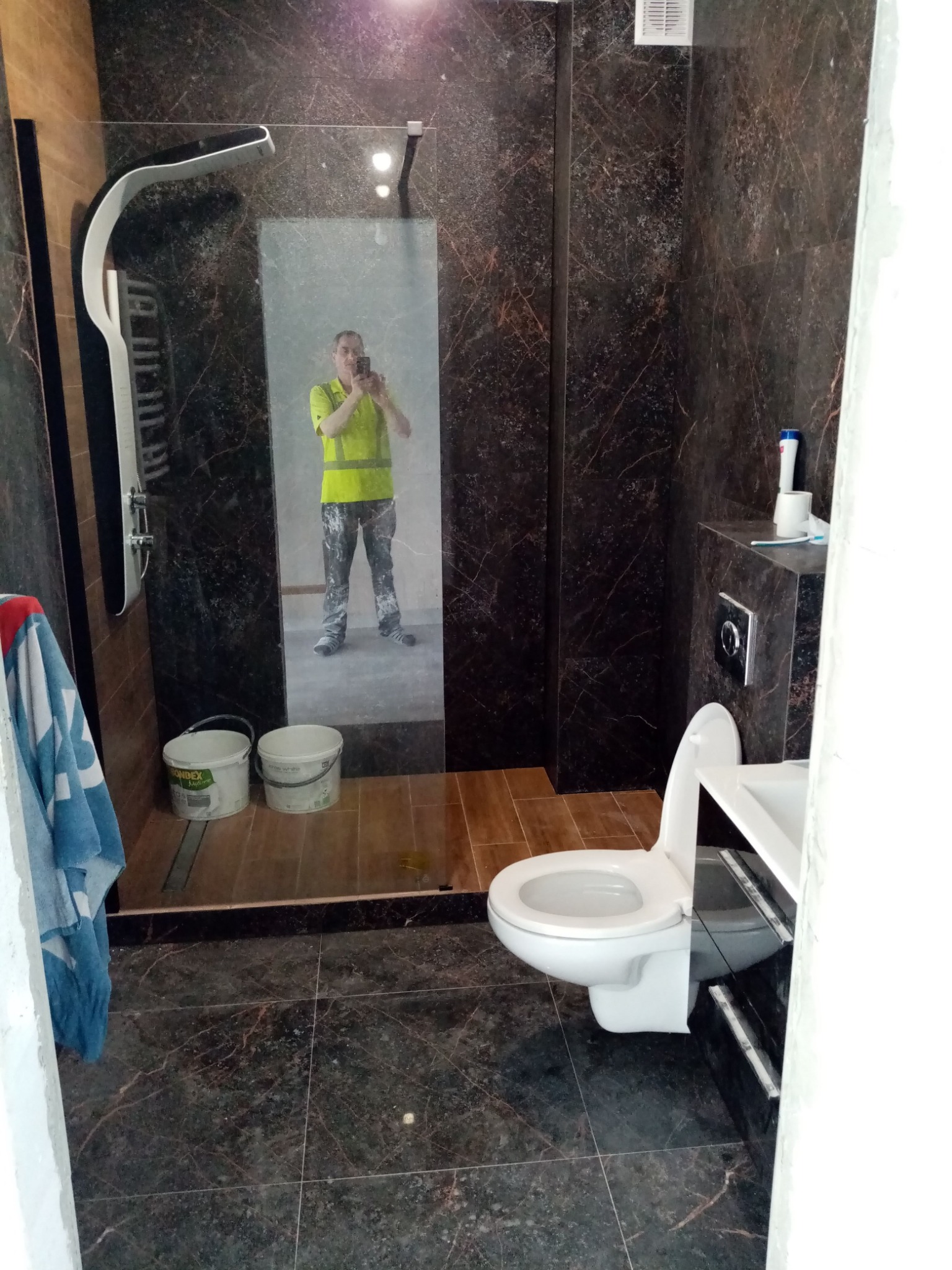 Ciemna łazienka z kabiną prysznicową typu walk-in, brązowe płytki imitujące marmur, widoczne dwa białe wiadra z farbą i odbicie pracownika w odblaskowej kamizelce robiącego zdjęcie.