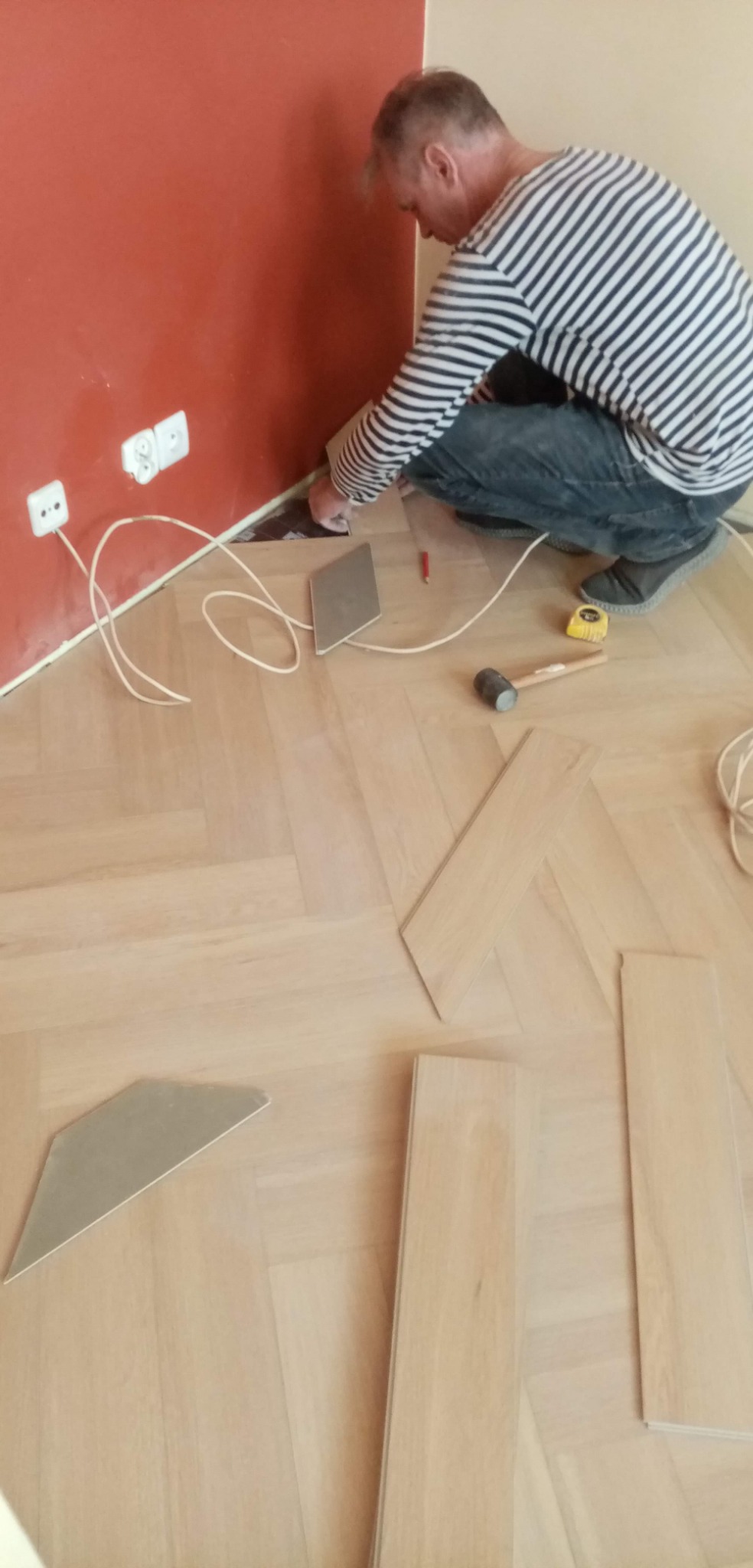 Parkiet układany w jodełkę, fachowiec w bluzie w paski docina listwę przypodłogową przy ścianie pomalowanej na dwa kolory, widoczne narzędzia: młotek, miarka, ołówek i docięte fragmenty paneli.