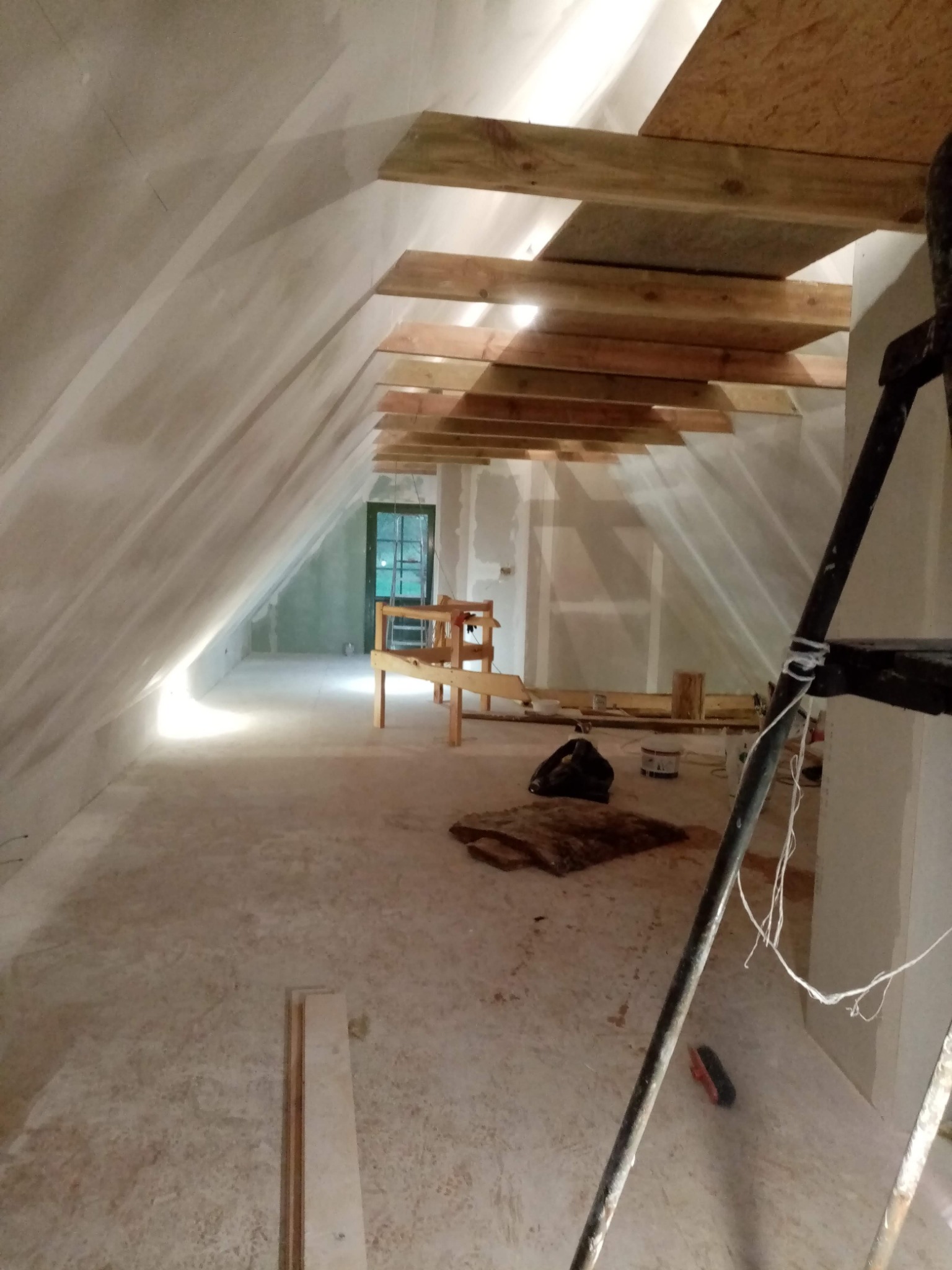 Poddasze w trakcie prac wykończeniowych: szpachlowane ściany, drewniana konstrukcja sufitu, drabina malarska w prawym dolnym rogu, okno w głębi pomieszczenia.