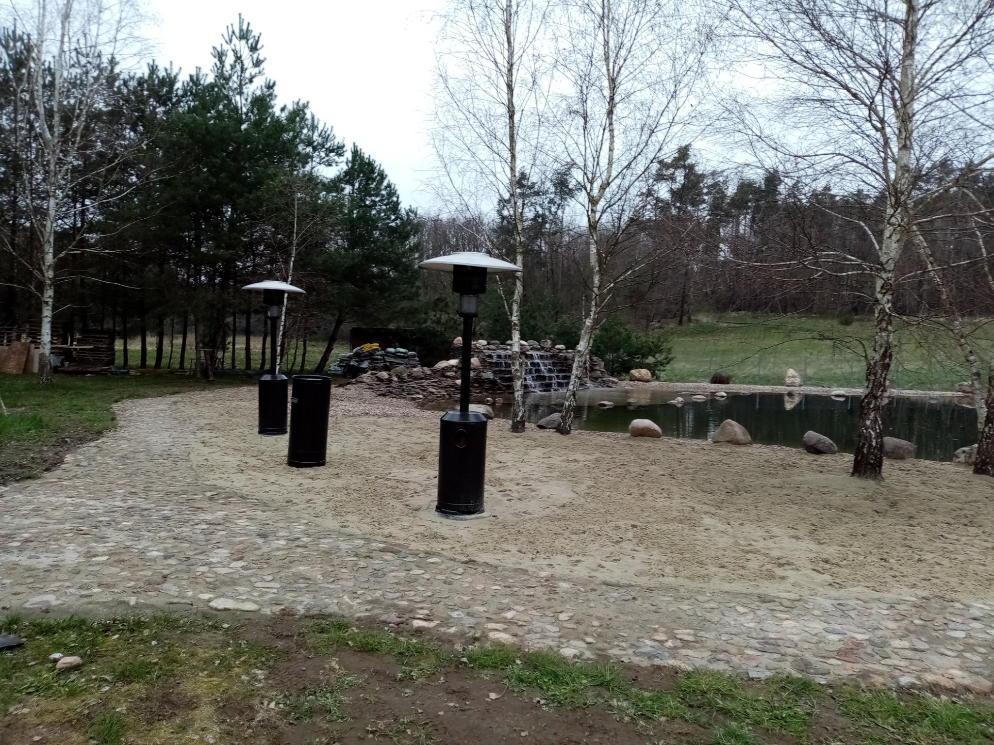 Ułożona z kamieni ścieżka prowadzi do małego stawu z wodospadem, oświetlona trzema czarnymi lampami grzewczymi z okrągłymi kloszami, otoczona brzozami i iglakami.