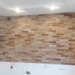 Daniel Kożuch Wykończenia wnętrz - Ściana wyłożona ceglastymi płytkami w odcieniach beżu i brązu, z widoczną wentylacją i gniazdami elektrycznymi na dole, podświetlona dwoma punktowymi lampami sufitowymi.