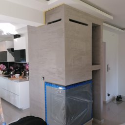 Daniel Kożuch Wykończenia wnętrz - Wnętrze w trakcie wykańczania: obudowa kominka z szarego tynku strukturalnego, zabezpieczona folią, widoczne otwory wentylacyjne i półki, kuchnia w tle, ubrania robocze na podłodze.