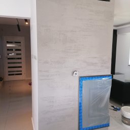 Daniel Kożuch Wykończenia wnętrz - Ściana z efektem betonu architektonicznego w jasnych odcieniach szarości, zabezpieczone okno folią malarską z niebieską taśmą, widoczny otwarty kontakt elektryczny, wiadro z farbą i narzędzia...