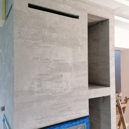 Daniel Kożuch Wykończenia wnętrz - Ściana z dekoracyjną, szarą strukturą tynku, z wnękami i otworem, widoczny fragment drabiny malarskiej w tle.