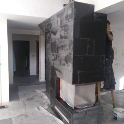 Daniel Kożuch Wykończenia wnętrz - Montaż obudowy kominka z czarnych płytek ceramicznych w trakcie prac wykończeniowych, widoczny glazurnik na drabinie, zabezpieczony wkład kominkowy folią.