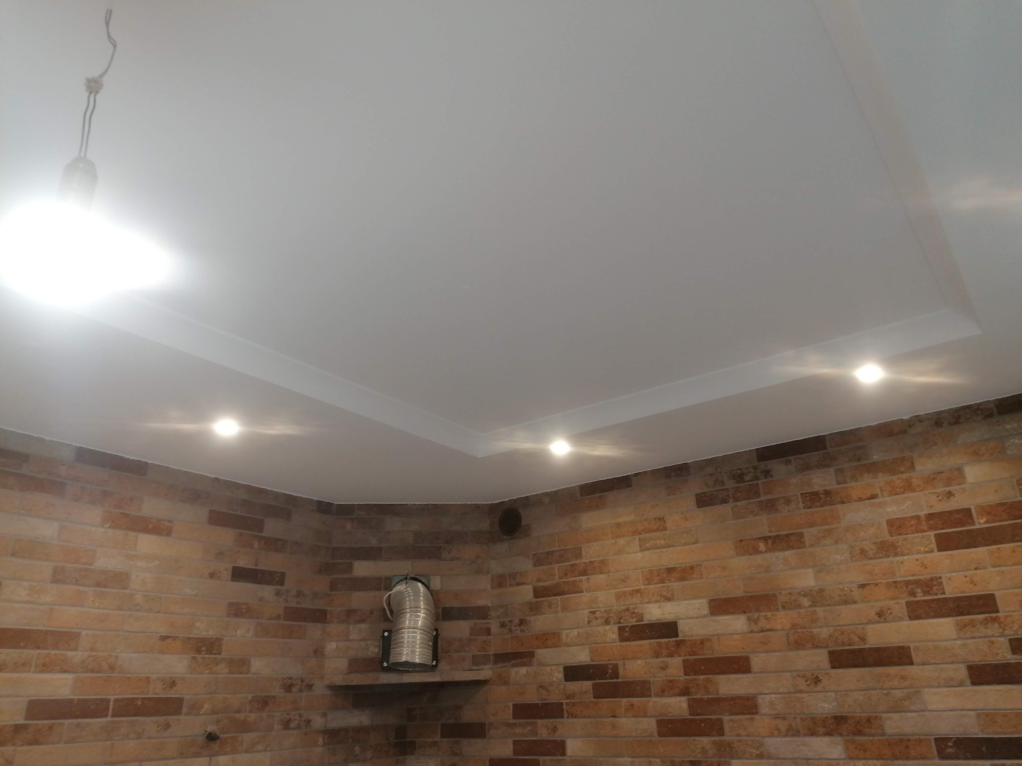 Biały sufit napinany z wbudowanymi punktowymi lampami LED, widoczny fragment ceglanej ściany z metalową rurą wentylacyjną