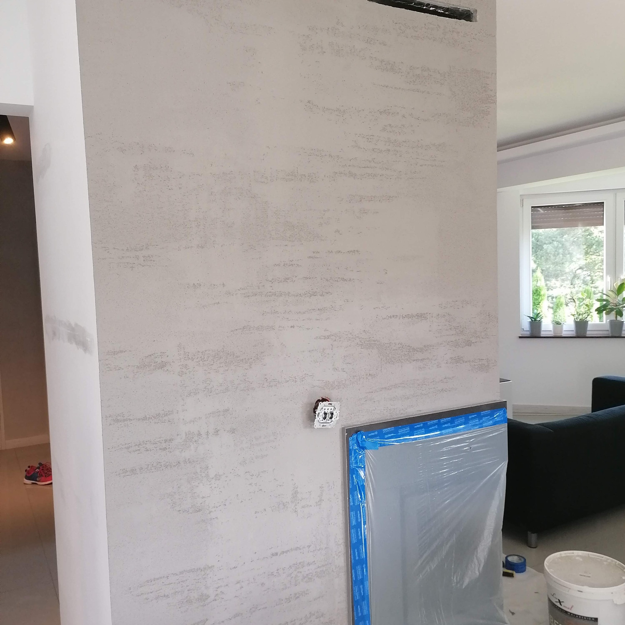 Ściana z dekoracyjną szarą fakturą, zabezpieczone folią drzwi, wystający przełącznik elektryczny, widoczny fragment wnętrza z oknem i roślinami.