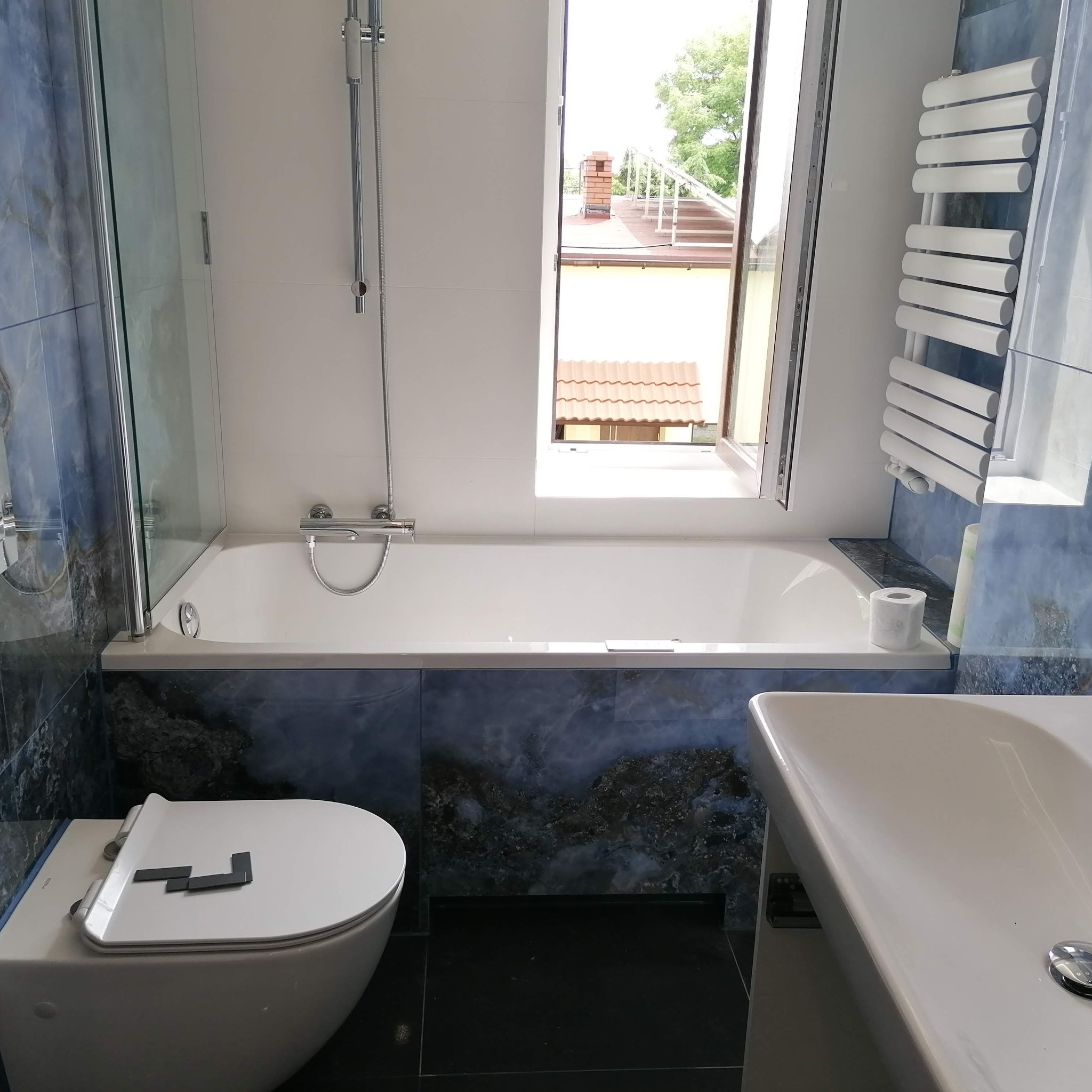 Wykończona łazienka z płytkami imitującymi marmur w odcieniach niebieskiego i szarości, biała wanna z parawanem nawannowym, grzejnik drabinkowy i umywalka z blatem, widok z okna na dach budynku.