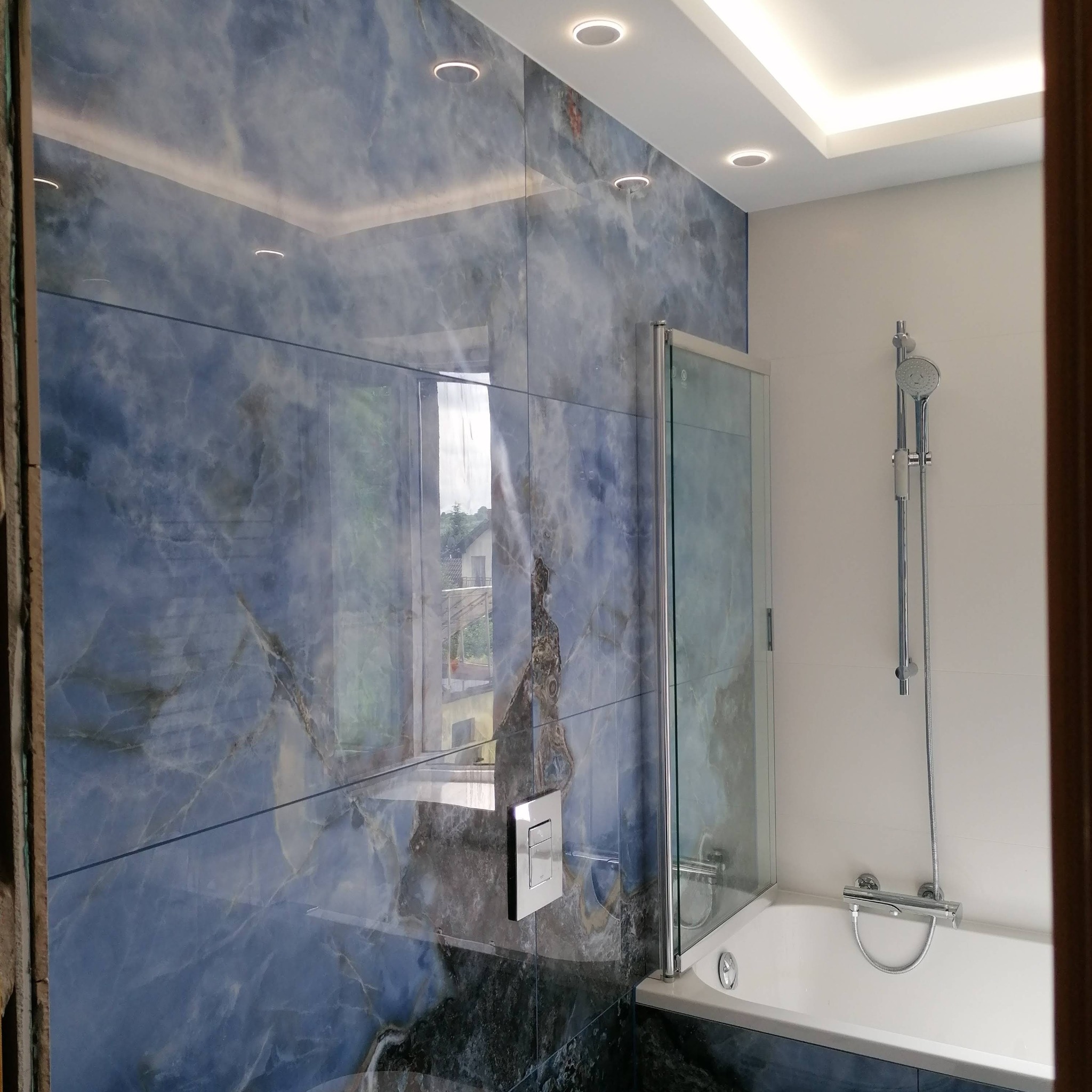Łazienka z wanną i kabiną prysznicową, ściany wyłożone niebiesko-szarymi płytkami imitującymi marmur, biała armatura, sufit z oświetleniem LED.