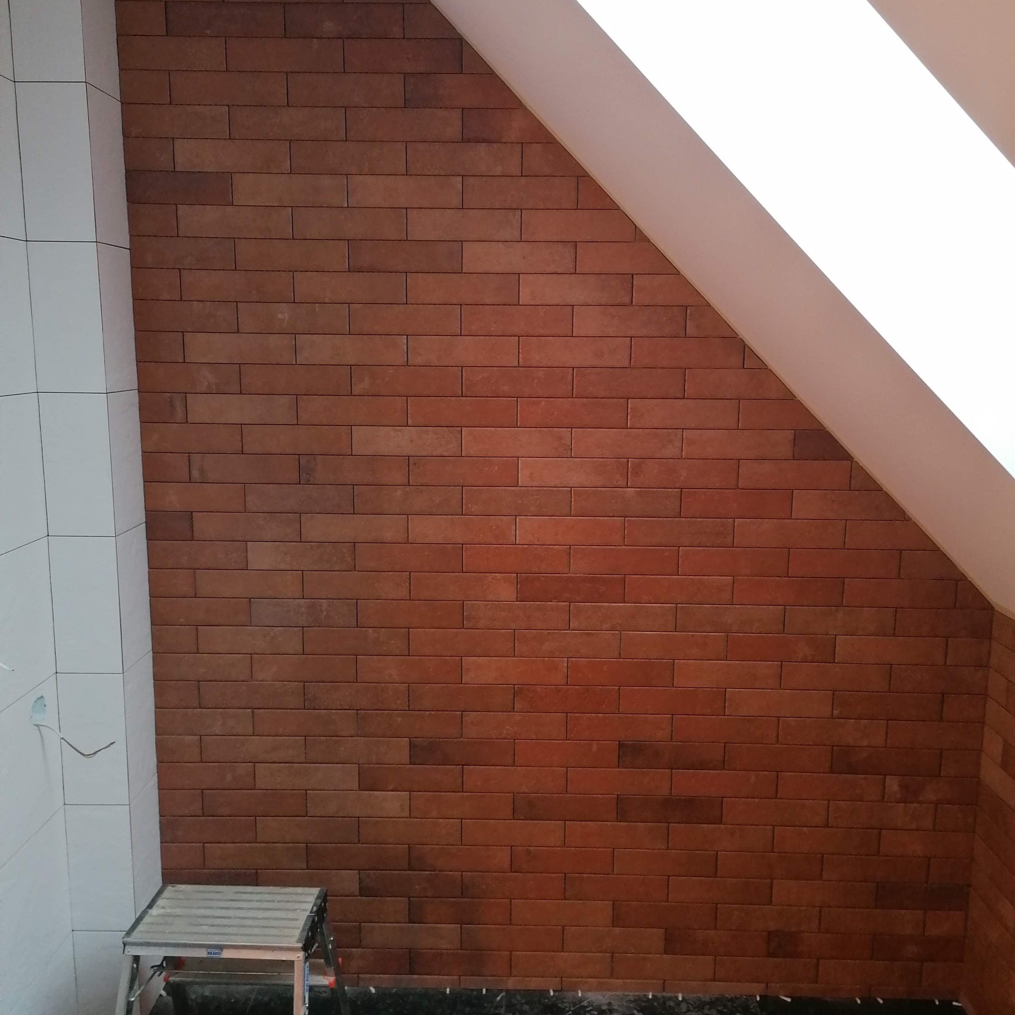 Ceglana okładzina ścienna w odcieniach brązu, ułożona w poziomym wzorze cegieł, kontrastująca z białą ścianą i skośnym sufitem na poddaszu, widoczna drabina malarska.